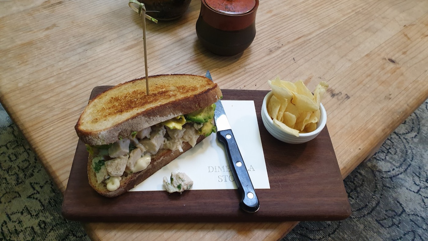 A toasted chicken and avocado sandwich on a wooden board, held with a skewer, with a small cup of chips and a steak knife