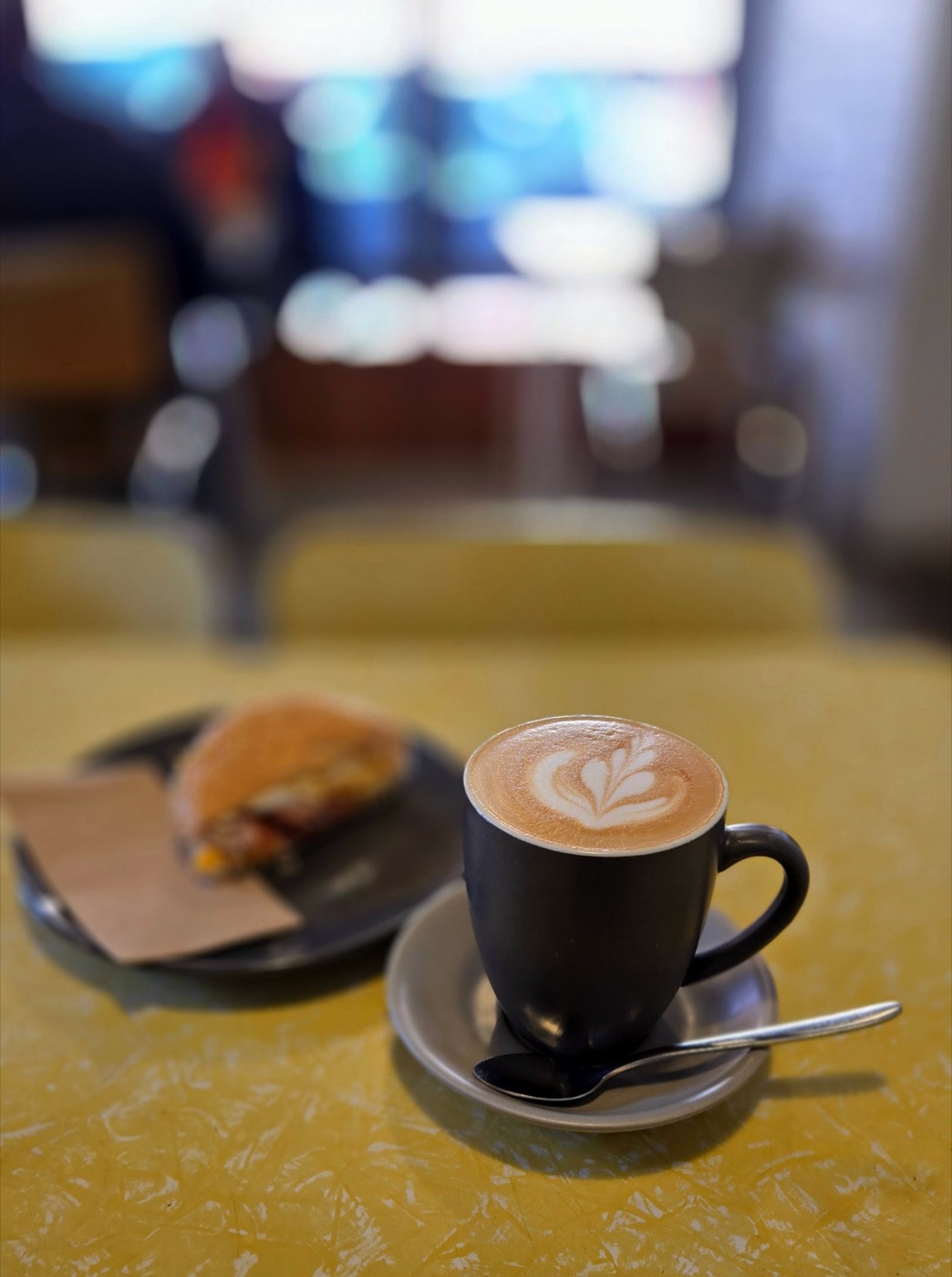 A flat white with latte art in a black cup on a yellow laminex table, with a toasted bun on a plate beside it