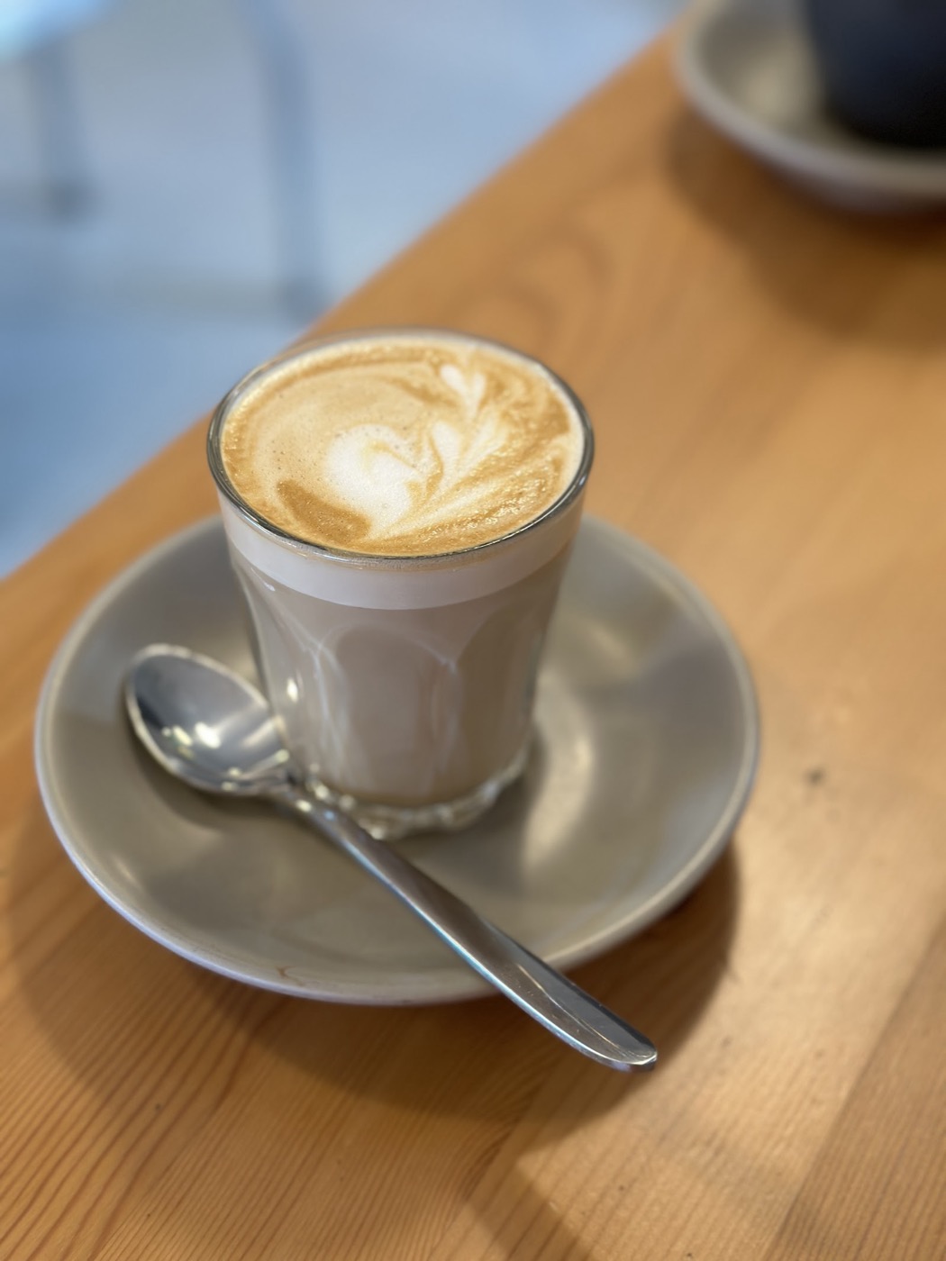 A latte in a glass with leaf latte art, on a saucer with a spoon, on a timber bench