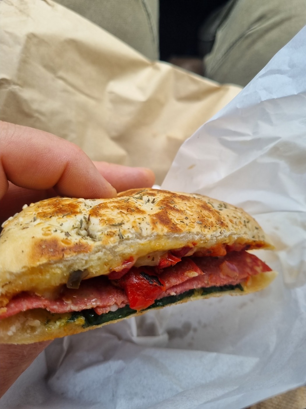 A toasted seeded roll held in a hand, filled with slices of salami, roasted red capsicum and greens