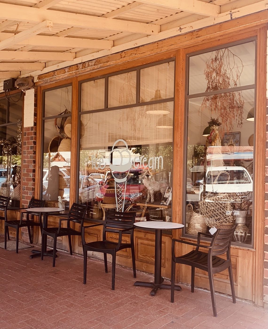 The shopfront under a timber verandah, with large glass windows, brick pillars and small cafe tables and chairs on the footpath