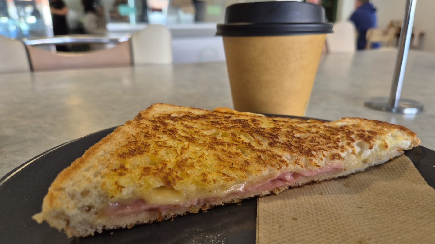 A golden toasted sandwich with a ham and cheese filling on a dark plate, with a takeaway coffee cup behind it