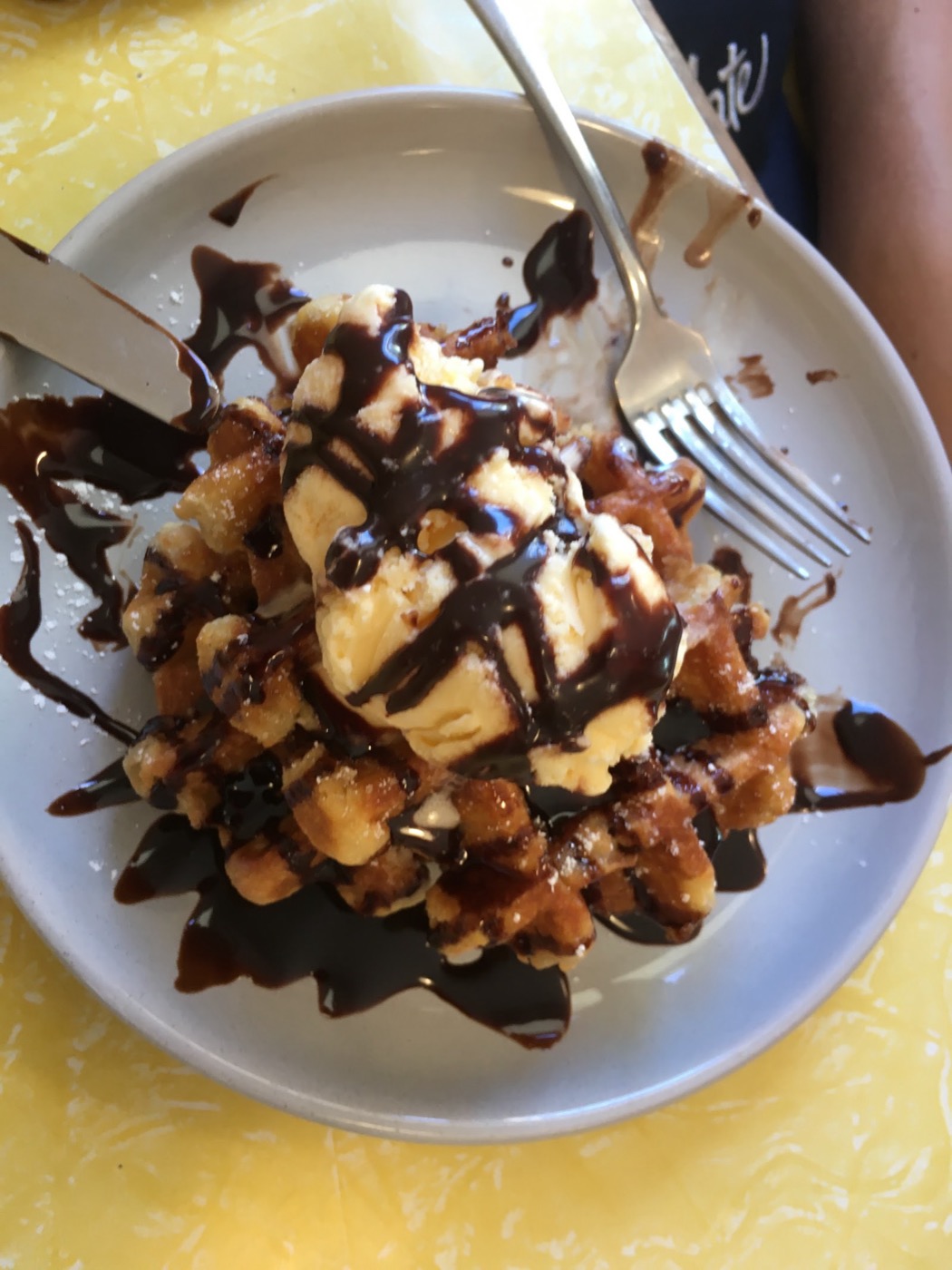 A waffle on a white plate topped with ice cream, nuts and chocolate sauce, on a yellow table